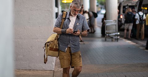 Man is on his mobile phone in an airport, wearing short-sleeved shirt and shorts.